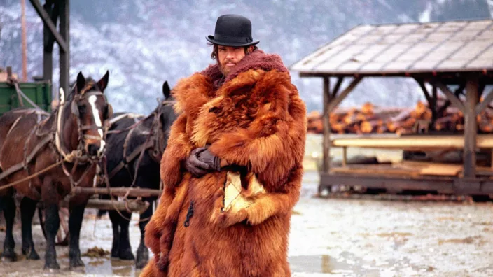 A man in a fur coat and bowler hat standing next to horses