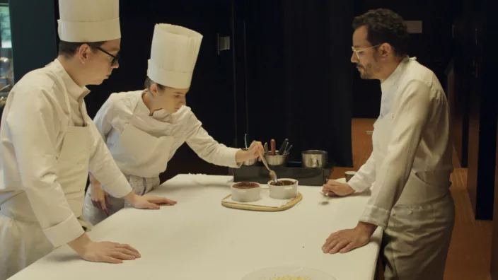 3 chefs dressed in whites, stand around a table testing a soufflé