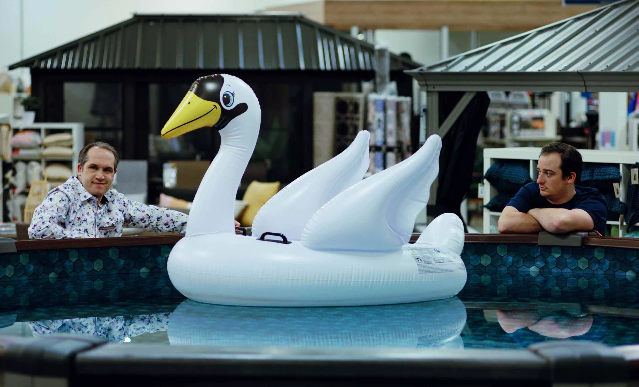 Two men beside a pool with an inflatable swan floating between them.
