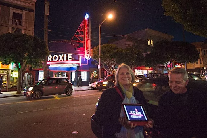 Two light skinned people stand across the street holding an ipad with a picture of the Roxie marquee. The partially lit Roxie marquee neon glows in the background.