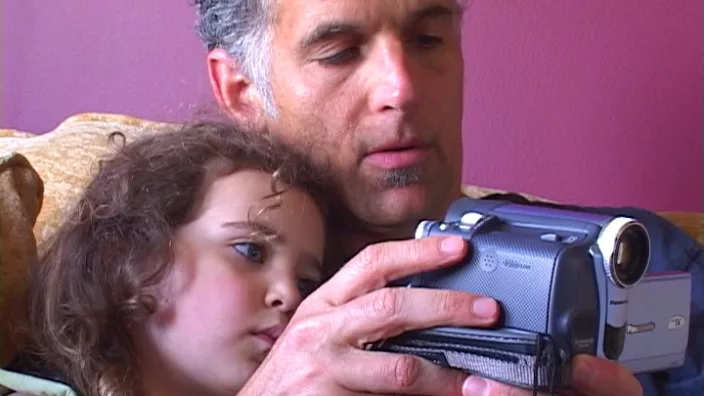 A man with greying hair with a young girl in his arms both inspect a movie camera his is holding.