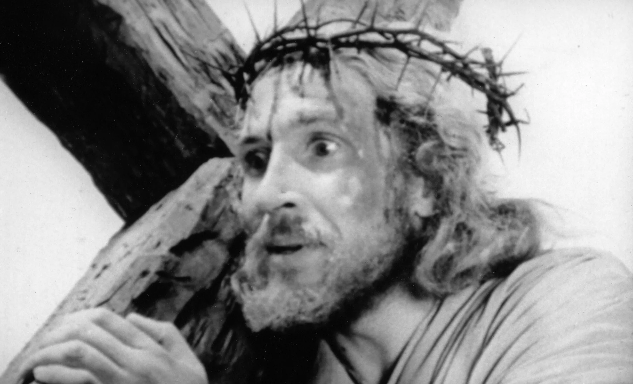 A black & white image of a man with long hair and a beard, wearing a crown of thorns, shouldering a large wooden cross.