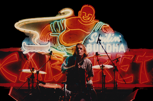 Glowing neon Buddha behind a musician. Mic stands and person with long hair and a stripe shirt and drums in front of the neon sign.