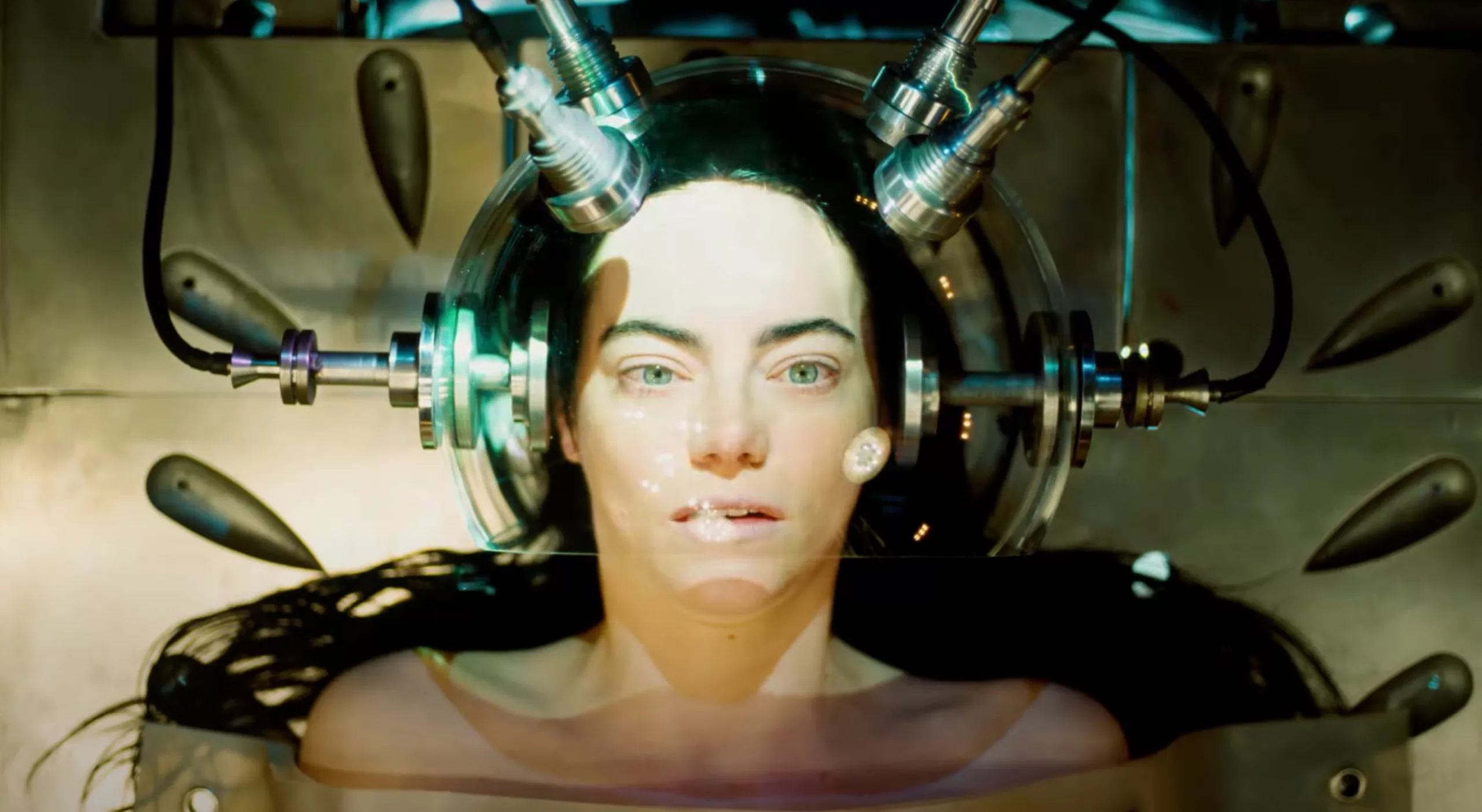 A woman looks up, eyes wide open, with her head encased in some sort of steam punk contraption on a table possibly in an operating room.