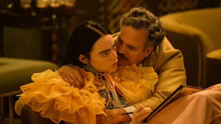 A young woman in an orange dress with a frilly collar is held close by a man with a mustache.