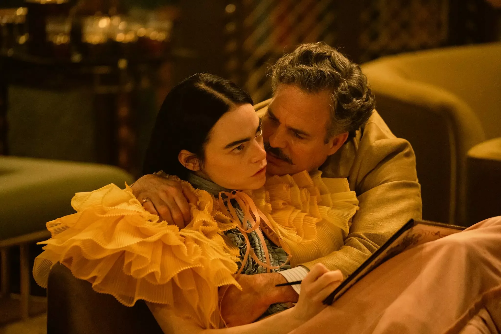 A young woman in an orange dress with a frilly collar is held close by a man with a mustache.