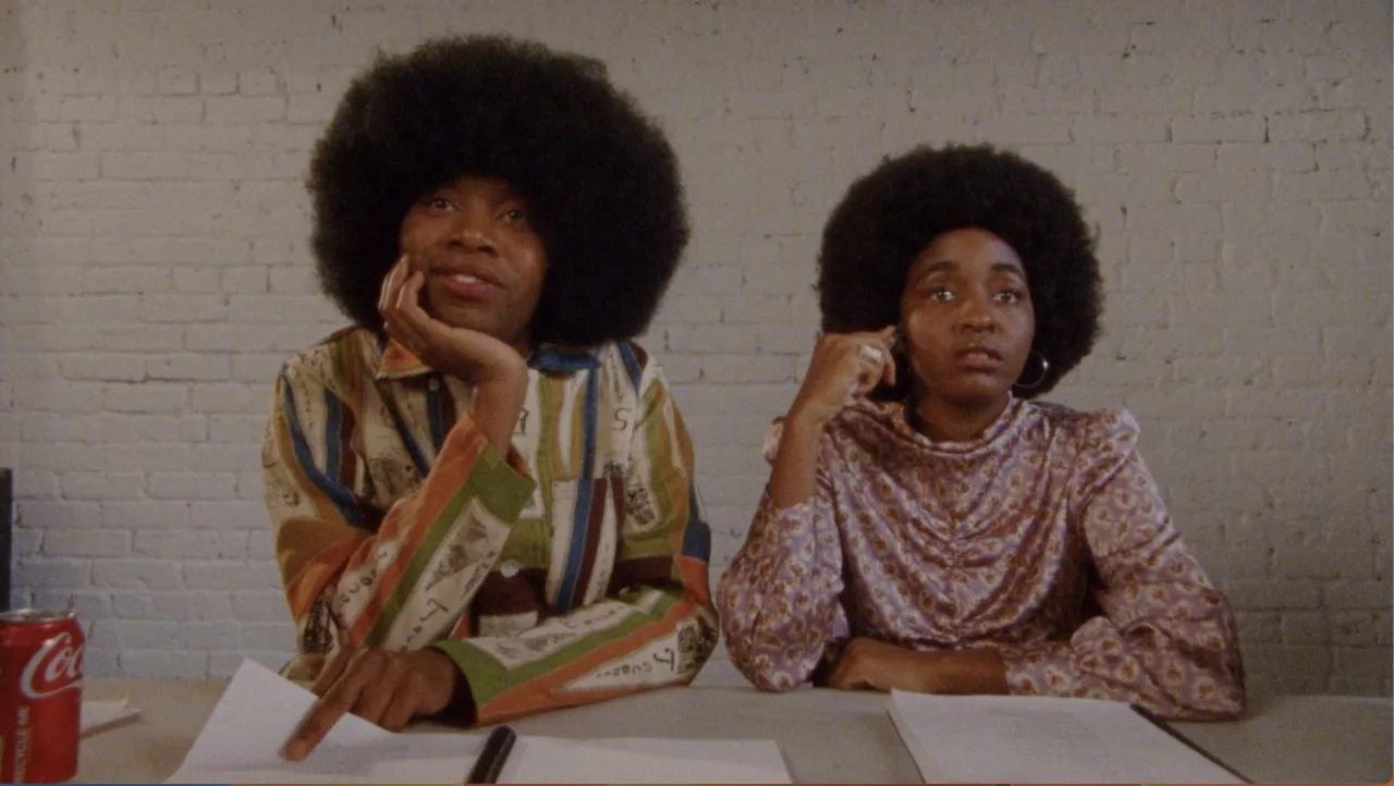 A Black man with an afro sits next to Black woman with an afro at a desk with a script
