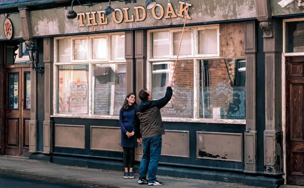 A man washes the window of a bar with the words THE OLD OAK on the side of the building, as a woman watches.