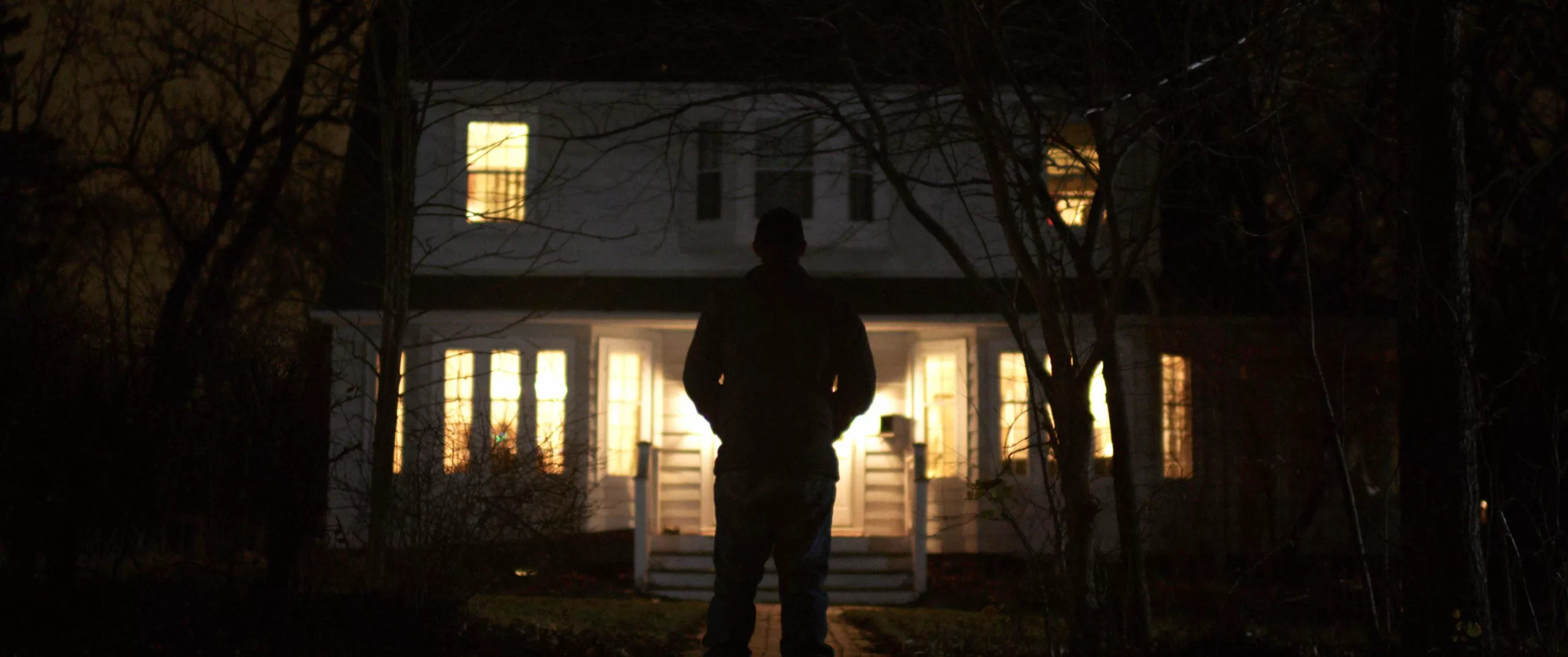 A scary figure with their back turned to us is silhouetted by lights inside a house in the background at night.