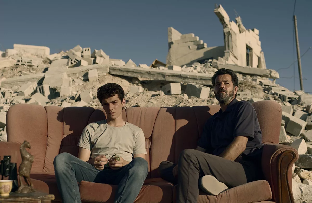 2 young men sit on a couch with the ruins of a concrete building in the background