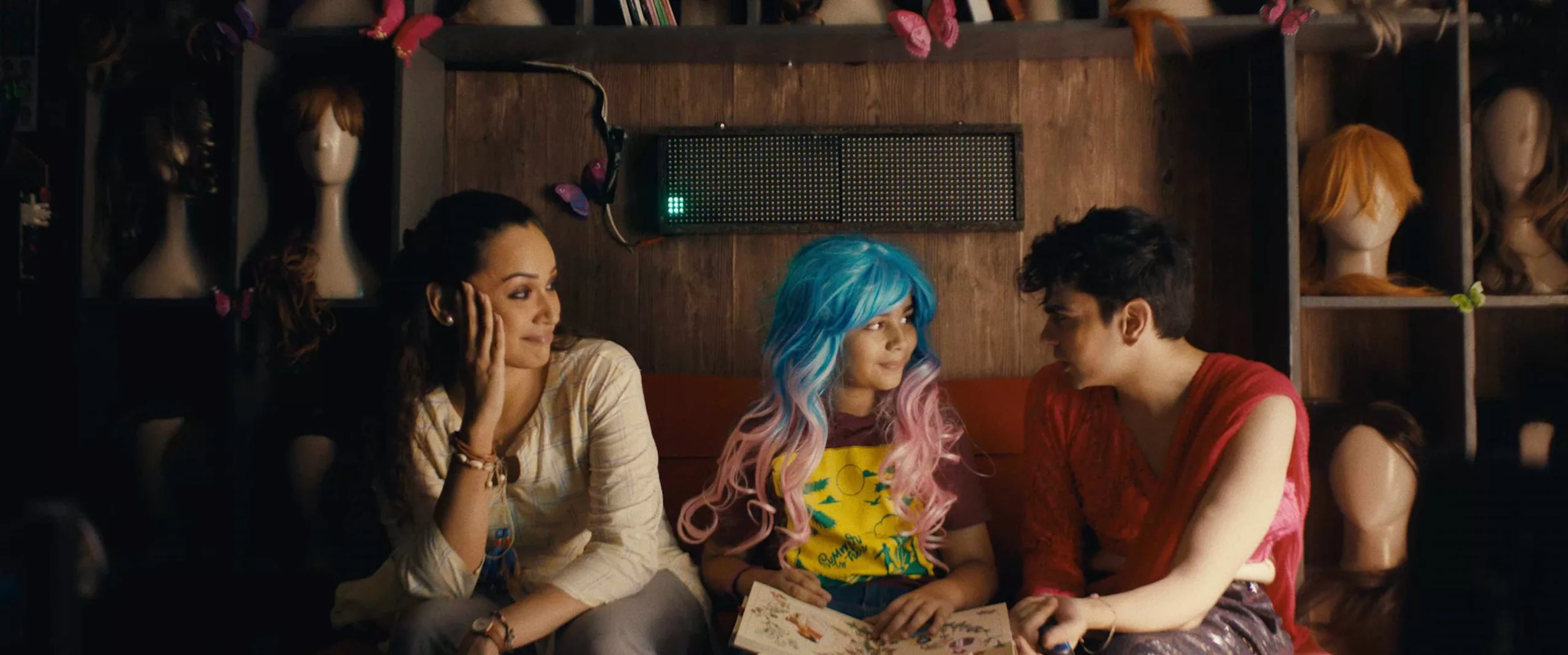 A young chilld in blue and pink wig sitting between two adults in front of a wall with other wigs
