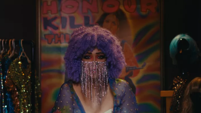 A woman with a purple wig & dress and a beaded face mask stares with an expression of urgency