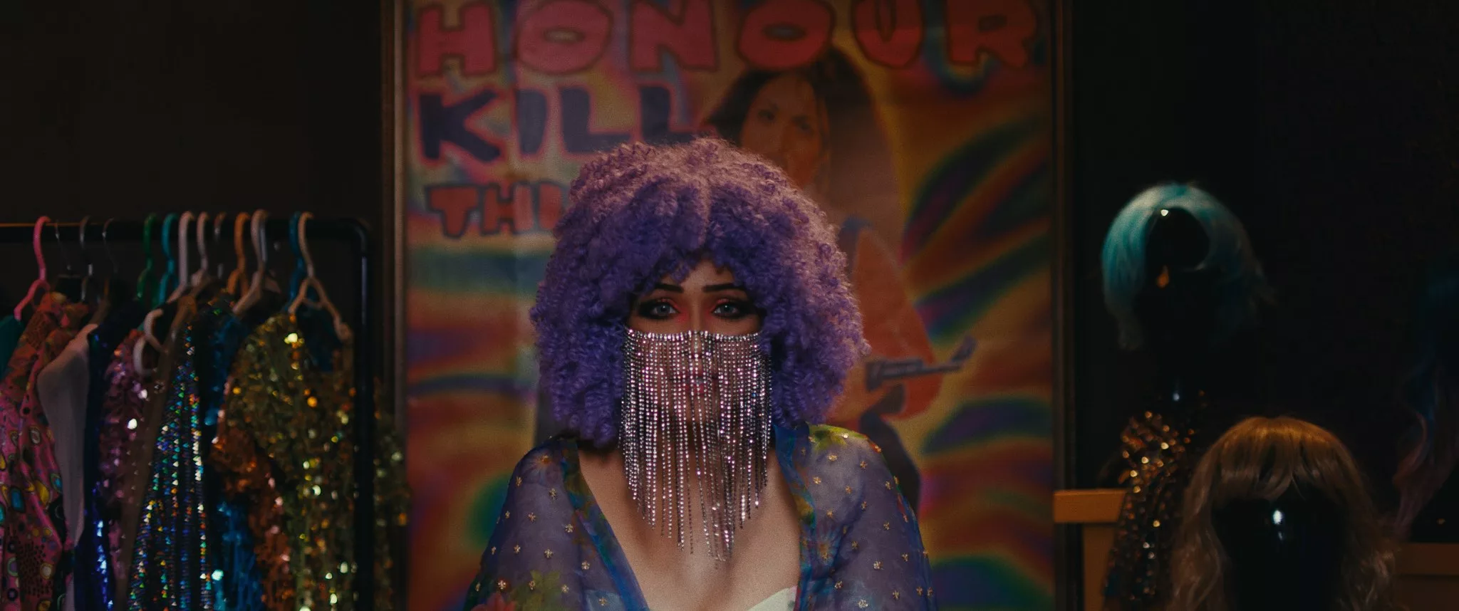 A woman with a purple wig & dress and a beaded face mask stares with an expression of urgency