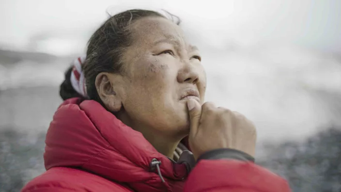 A Nepali woman in a red jacket holds her chin an pensively stars upward