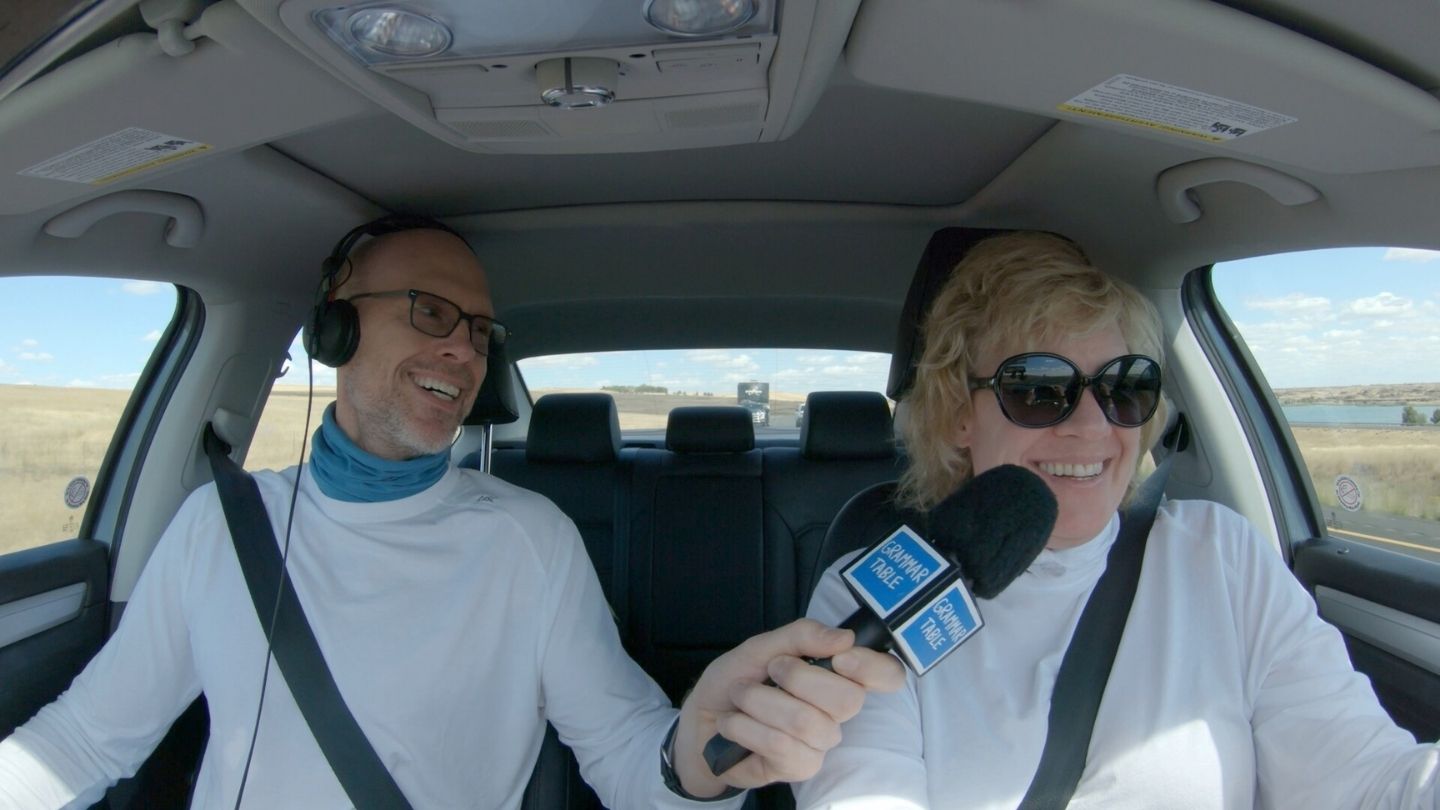 man in white sweater and a mic next to a woman in a car
